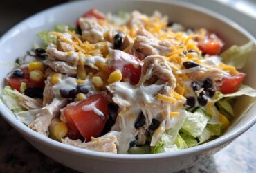 A close-up of a white bowl filled with a vibrant southwest salad featuring shredded chicken, black beans, corn, tomatoes, and creamy dressing.