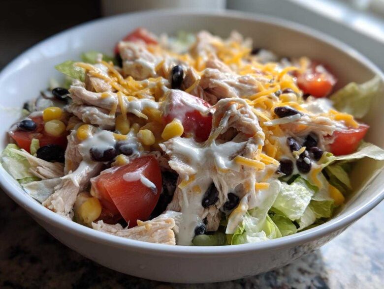 A close-up of a white bowl filled with a vibrant southwest salad featuring shredded chicken, black beans, corn, tomatoes, and creamy dressing.