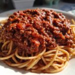 Close-up of spaghetti topped generously with rich, meaty bolognese sauce on a white plate.