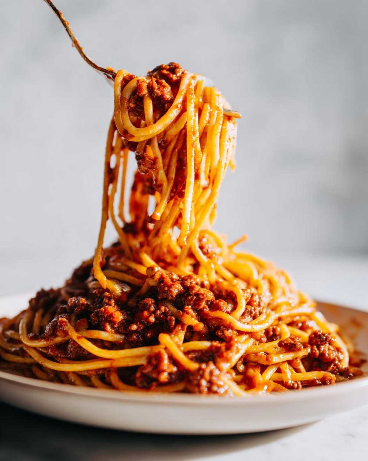 A fork lifts spaghetti heavily coated in rich, meaty bolognese sauce from a plate.