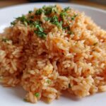 Close-up of a mound of perfectly cooked, reddish-orange Spanish rice garnished with fresh chopped parsley on a white plate.