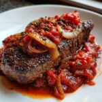 Close-up of a perfectly seared steak pizzaiola covered in rich tomato sauce, sautéed onions, and herbs.