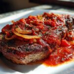 A perfectly seared steak pizzaiola topped with rich tomato sauce, sautéed onions, and herbs, served on a white plate.