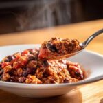 A spoonful of steaming, rich high protein chili, featuring ground meat and kidney beans, lifted from a white bowl.