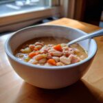 Close-up of a steaming white bowl filled with hearty bean soup containing white beans, carrots, celery, and ham chunks.