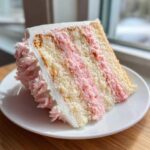 A tall, layered slice of Strawberry Whipped Cream Cake showing vanilla sponge layers and pink strawberry filling on a white plate.