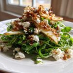 A vibrant pear salad featuring arugula, crumbled white cheese, and caramelized onions served on a white plate.