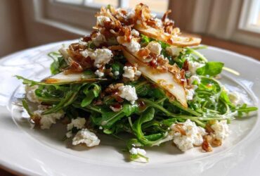 A vibrant pear salad featuring arugula, crumbled white cheese, and caramelized onions served on a white plate.
