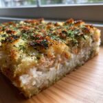 A thick, golden-brown slice of baked sushi bake featuring layers of white rice and creamy filling, topped with toasted seaweed flakes.