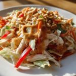 A vibrant plate of thai chicken salad featuring shredded chicken, cabbage, carrots, red pepper, and a rich peanut dressing topped with chopped peanuts.