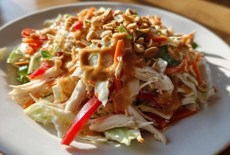 A vibrant plate of thai chicken salad featuring shredded chicken, cabbage, carrots, red pepper, and a rich peanut dressing topped with chopped peanuts.