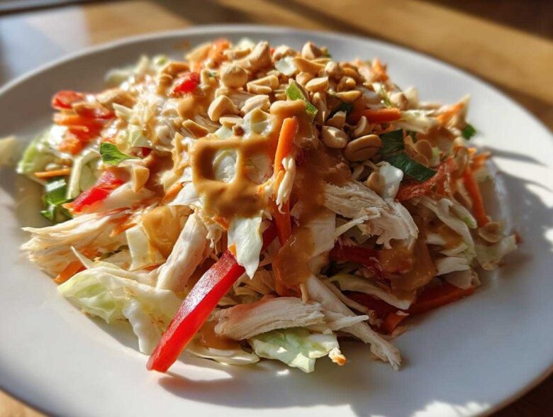 A vibrant plate of thai chicken salad featuring shredded chicken, cabbage, carrots, red pepper, and a rich peanut dressing topped with chopped peanuts.