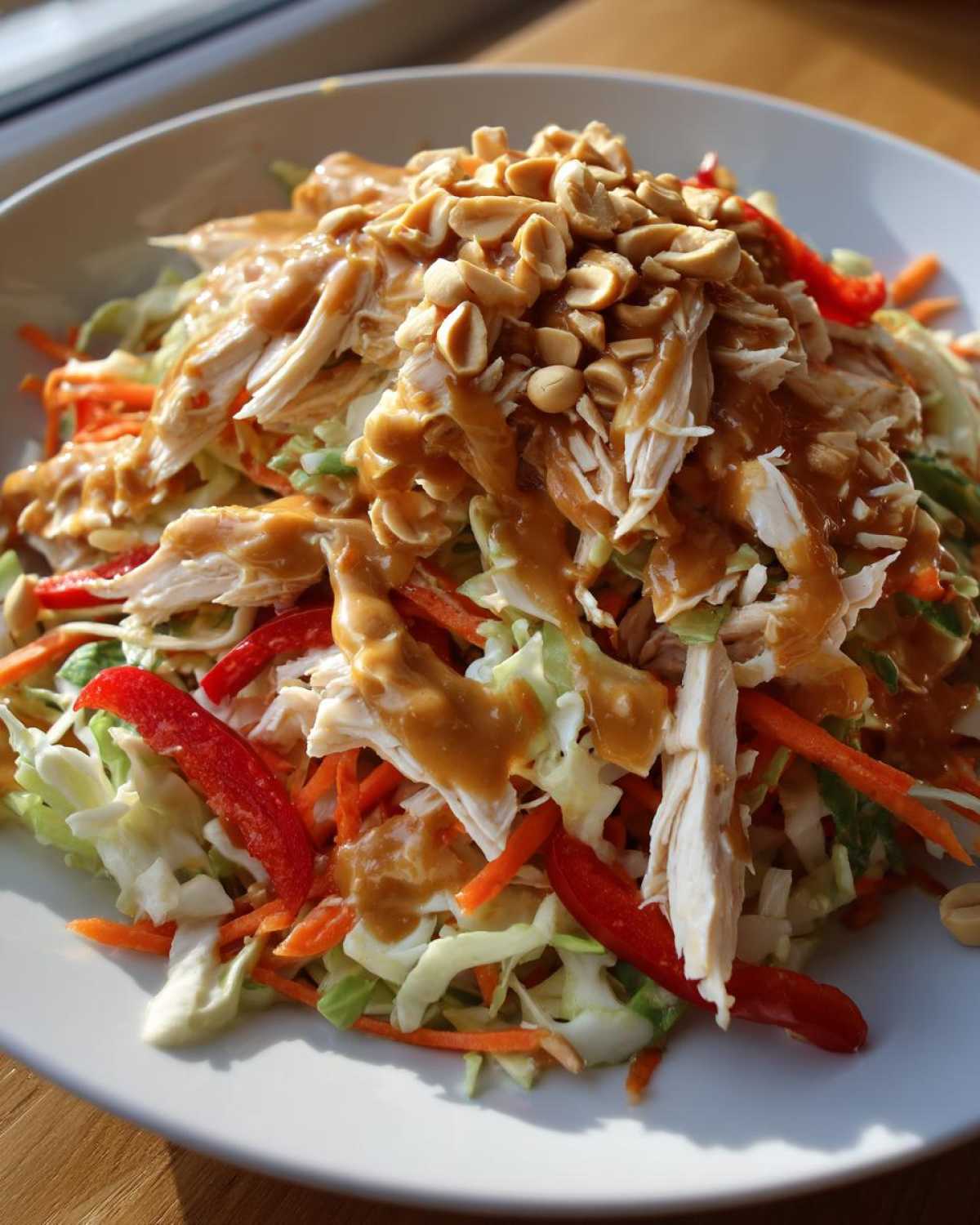 Close-up of a vibrant thai chicken salad featuring shredded chicken, cabbage, carrots, red peppers, and a generous drizzle of peanut sauce topped with crushed peanuts.