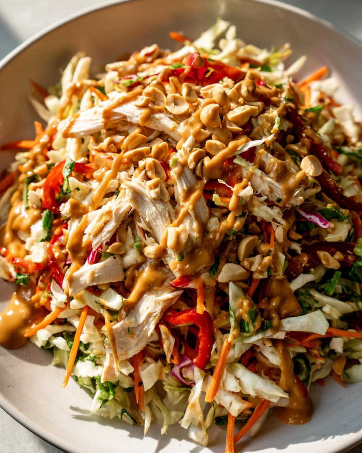 Close-up of a vibrant thai chicken salad featuring shredded chicken, cabbage, carrots, and a generous drizzle of peanut sauce and chopped peanuts.
