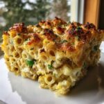 A close-up of a thick slice of creamy tuna pasta bake with a golden, crispy cheese topping and visible green peas.