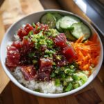 A vibrant, close-up view of a Tuna Poke Bowl featuring marinated raw tuna cubes over rice, topped with green onions and sesame seeds.