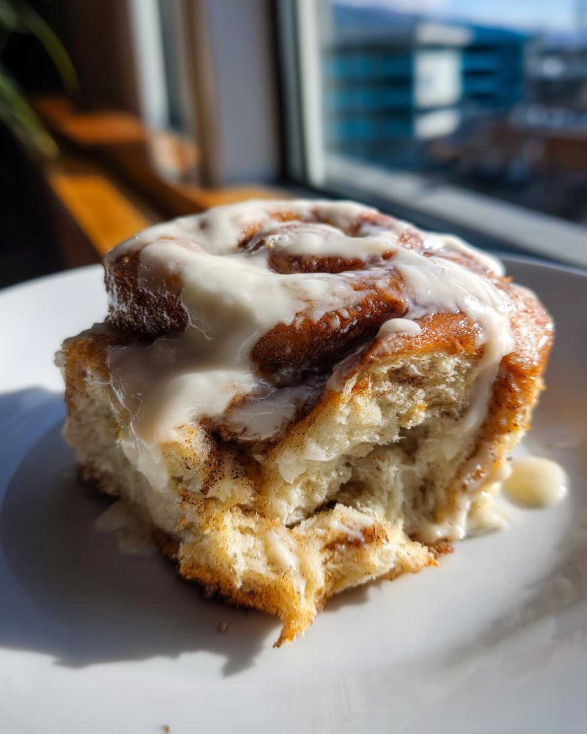 A close-up of one Ultimate Cinnamon Rolls with Creamy Glaze, heavily frosted and sitting on a white plate near a window.