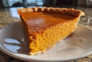 A close-up, sunlit slice of rich orange sweet potato pie resting on a white plate.