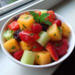 Close-up of a colorful Fruit Salad featuring cantaloupe, strawberries, raspberries, and blackberries, topped with fresh mint.