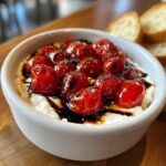 A close-up of a warm bruschetta dip featuring roasted cherry tomatoes over creamy cheese, drizzled with balsamic glaze, served with bread slices.