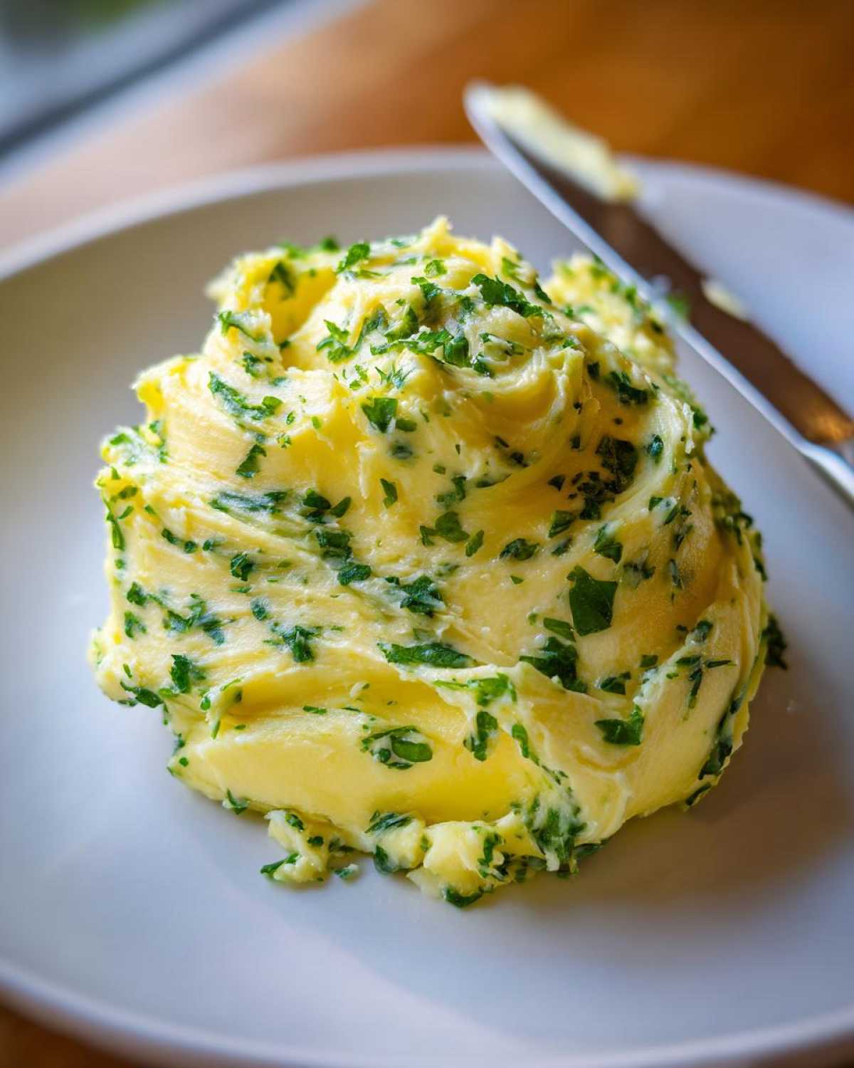 A mound of whipped, yellow garlic butter mixed with chopped green herbs, likely parsley, served on a white plate next to a knife.