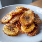 A pile of crispy Air Fryer Cinnamon Sugar Banana Chips coated heavily in sparkling sugar and cinnamon on a white plate.