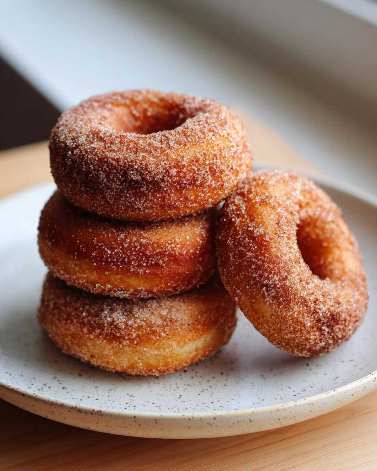 A close-up of four golden Baked Cinnamon Sugar Donuts stacked on a speckled white plate.