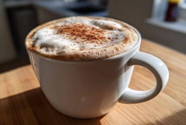 A close-up of a frothy Banana Bread Latte topped with cinnamon in a white mug, sitting on a wooden surface.