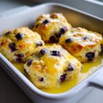 Four freshly baked Blueberry Butter Swim Biscuits soaking in a pool of bright yellow lemon glaze in a white baking dish.