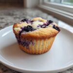 A single, golden-brown Blueberry Cream Cheese Muffin with a dollop of cream cheese filling on top, served on a white plate.