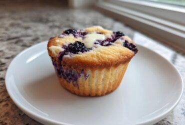 A single, golden-brown Blueberry Cream Cheese Muffin with a dollop of cream cheese filling on top, served on a white plate.