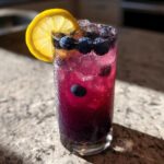 A tall, sweating glass filled with vibrant Blueberry Lemonade, ice, fresh blueberries, and a lemon slice garnish.