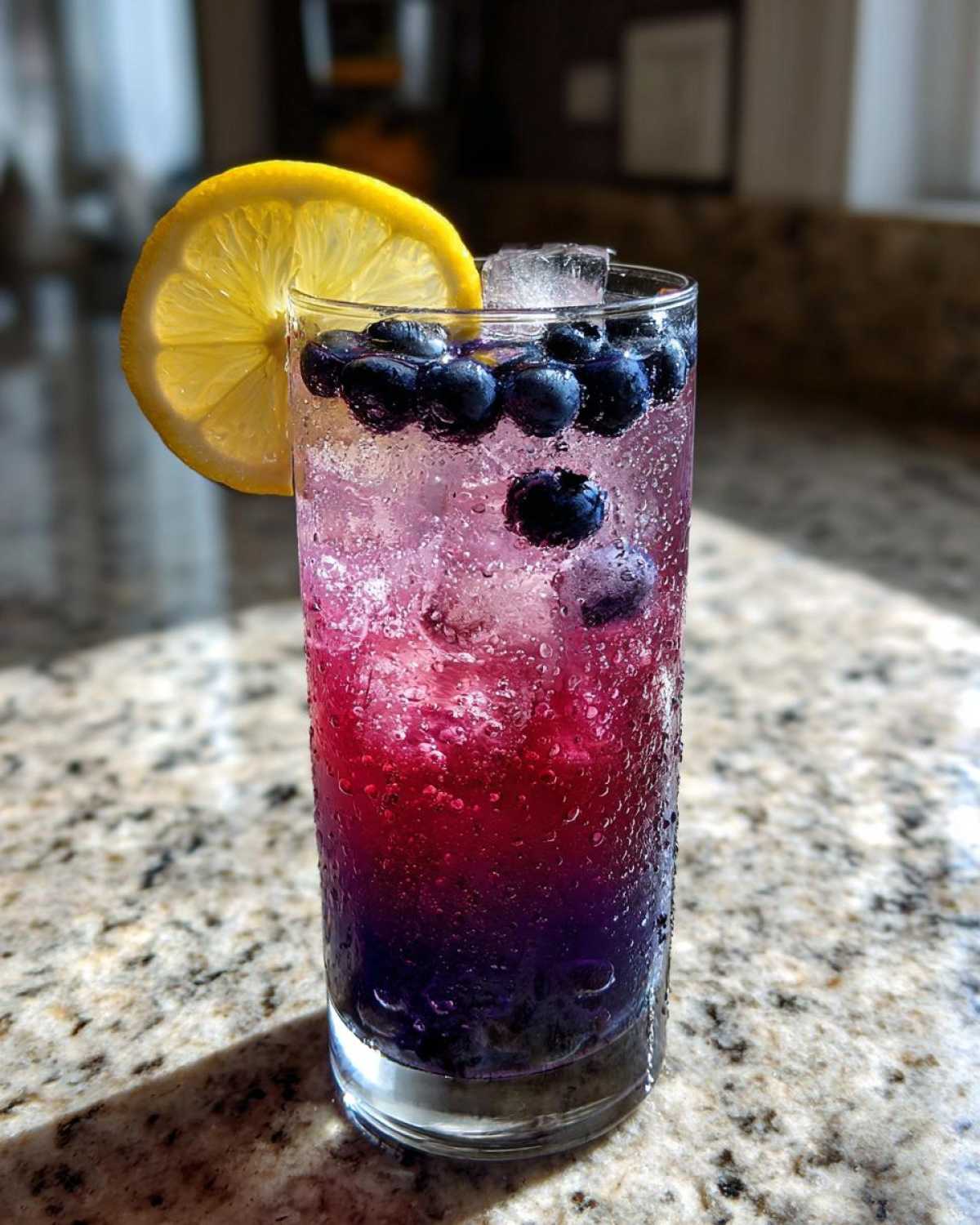 A tall glass of layered Blueberry Lemonade with ice, fresh blueberries, and a lemon slice garnish.