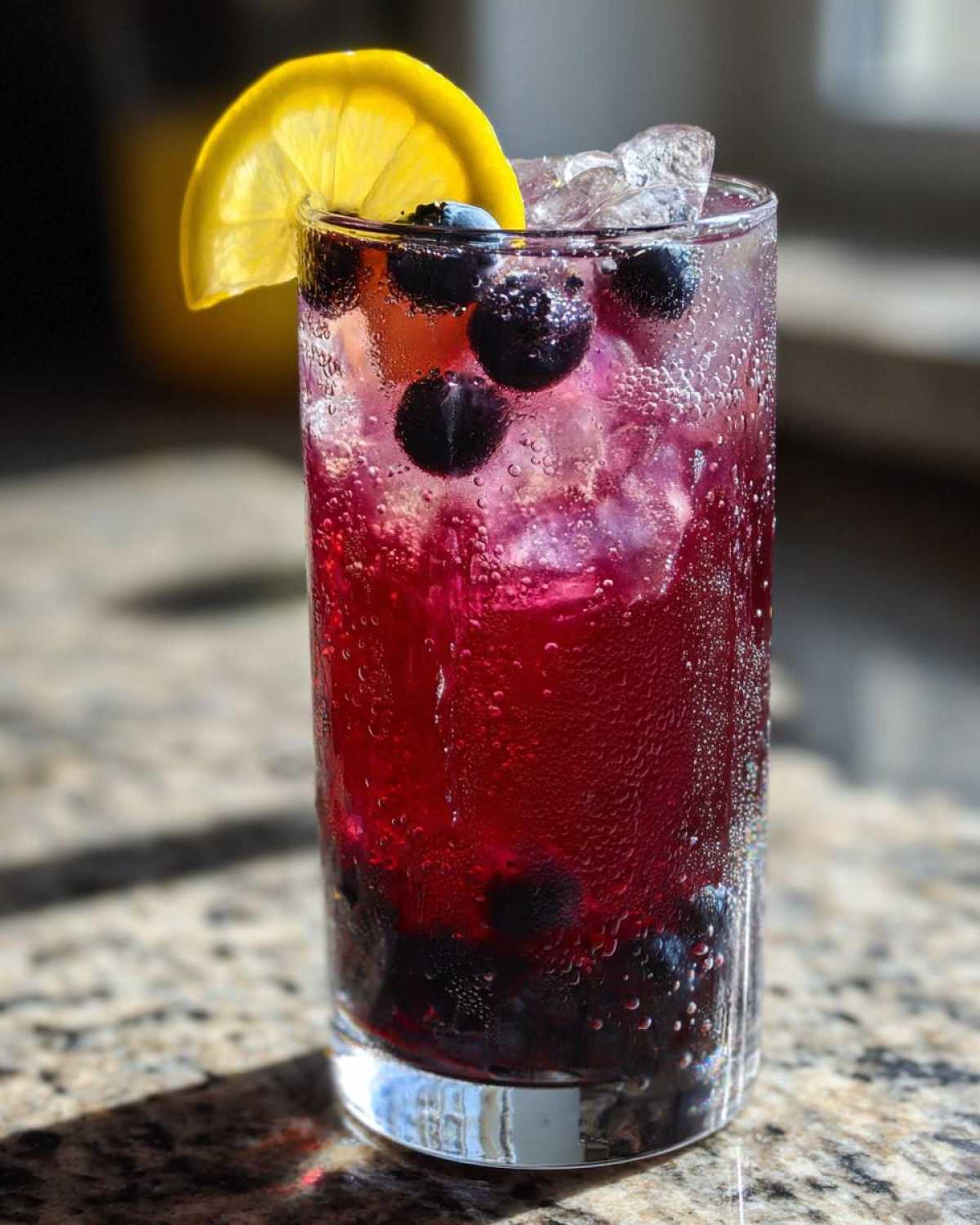 Close-up of a tall glass filled with vibrant Blueberry Lemonade, ice, fresh blueberries, and a lemon slice garnish.
