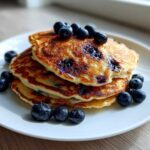A stack of three golden-brown Blueberry Pancakes topped and garnished with fresh blueberries on a white plate.