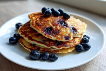 A stack of three golden-brown Blueberry Pancakes topped and garnished with fresh blueberries on a white plate.