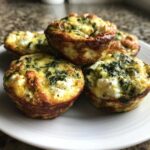 A close-up of several golden brown Breakfast Mini Quiches topped with spinach and white feta cheese on a white plate.