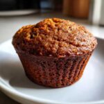 A close-up of a single, richly browned Brown Sugar Banana Muffin sitting on a white plate.