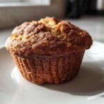 A single, golden-brown Brown Sugar Banana Muffin sitting on a white plate, highlighted by natural sunlight.