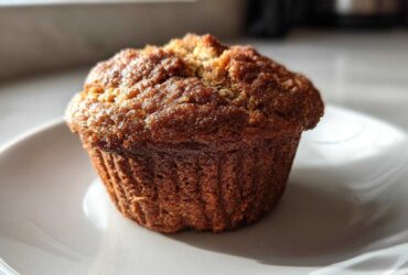 A single, golden-brown Brown Sugar Banana Muffin sitting on a white plate, highlighted by natural sunlight.