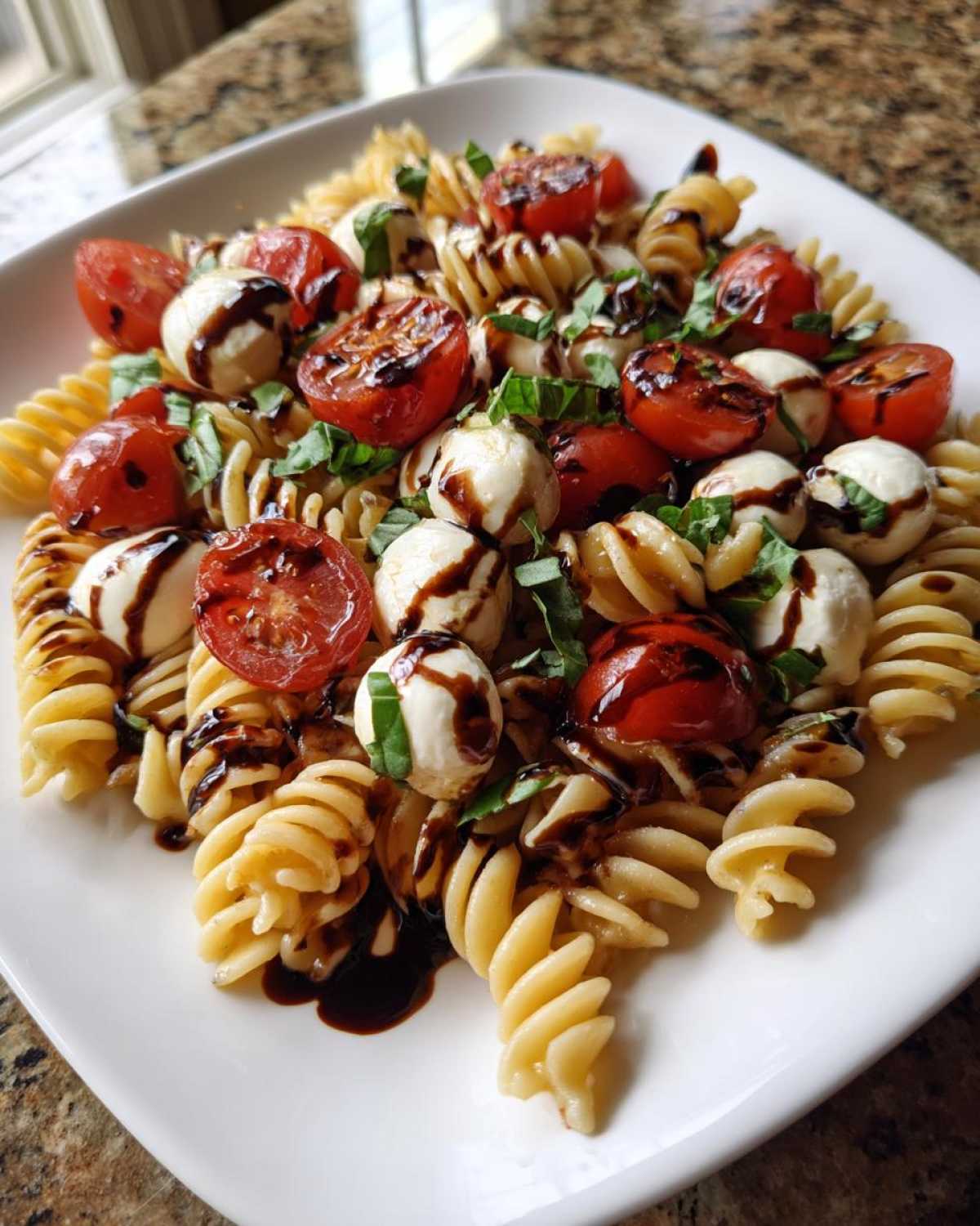 A close-up of a serving of Caprese Pasta Salad featuring rotini pasta, cherry tomatoes, mozzarella balls, and basil drizzled with balsamic glaze.