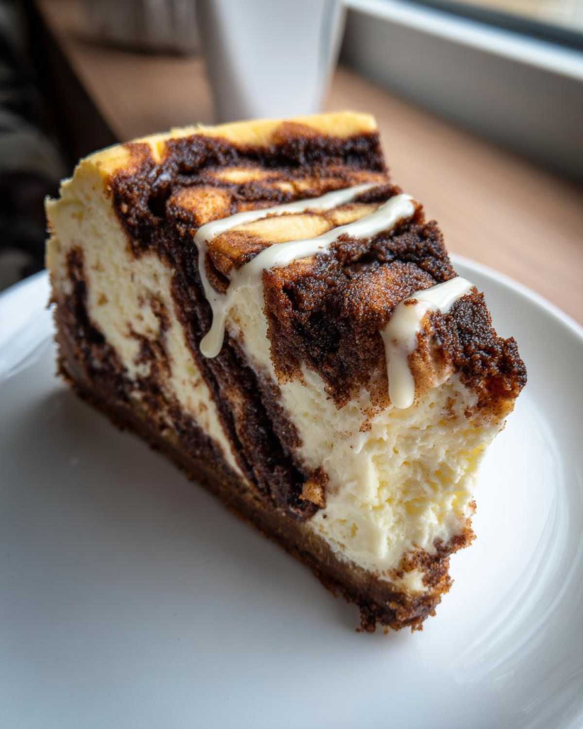 A close-up of a rich slice of Cinnamon Roll Cheesecake featuring a marbled cinnamon swirl and a white icing drizzle on a white plate.