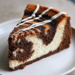 A close-up of a rich slice of Cinnamon Roll Cheesecake showing marbled dark chocolate and cream cheese layers, drizzled with white icing.