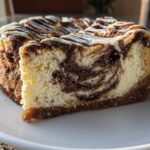 A close-up slice of rich Cinnamon Roll Cheesecake showing a beautiful cinnamon swirl pattern in the creamy filling, topped with white icing.