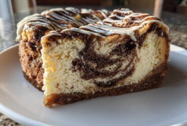 A close-up slice of rich Cinnamon Roll Cheesecake showing a beautiful cinnamon swirl pattern in the creamy filling, topped with white icing.