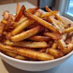 A close-up of a white bowl filled with golden brown, crispy air fryer french fries seasoned with salt and pepper.