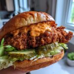 A hand holding a tall, perfectly assembled Crispy Chicken Sandwich featuring a thick fried patty, pickles, lettuce, and orange sauce on a brioche bun.