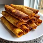 A stack of golden brown, crispy oven rolled chicken taquitos piled high on a white plate.