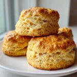 A stack of golden brown, tall, and flaky Cream Scones resting on a white plate near a window.