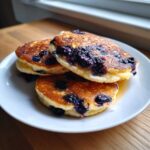 Close-up of three golden-brown Blueberry Pancakes stacked on a white plate, bursting with cooked blueberries.